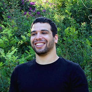 A man smiles while standing in front of dense green foliage with some purple flowers visible, wearing a black shirt and appearing relaxed and happy in a natural outdoor setting.