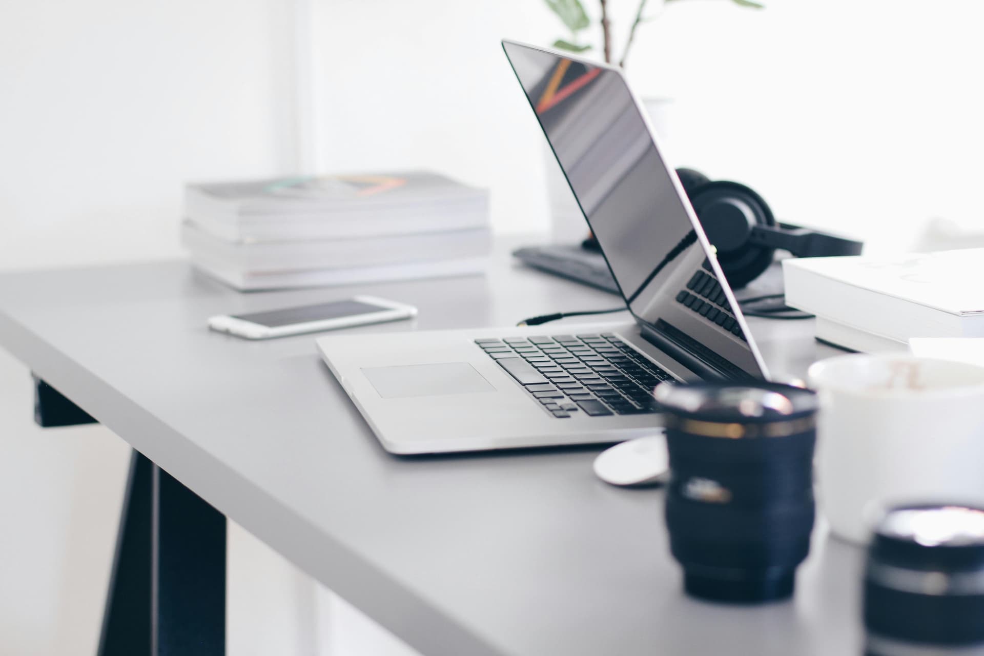 A laptop lies open on a modern, gray desk, accompanied by a smartphone, stacked books, headphones, a camera lens, and a coffee cup in a bright, minimalist office space.