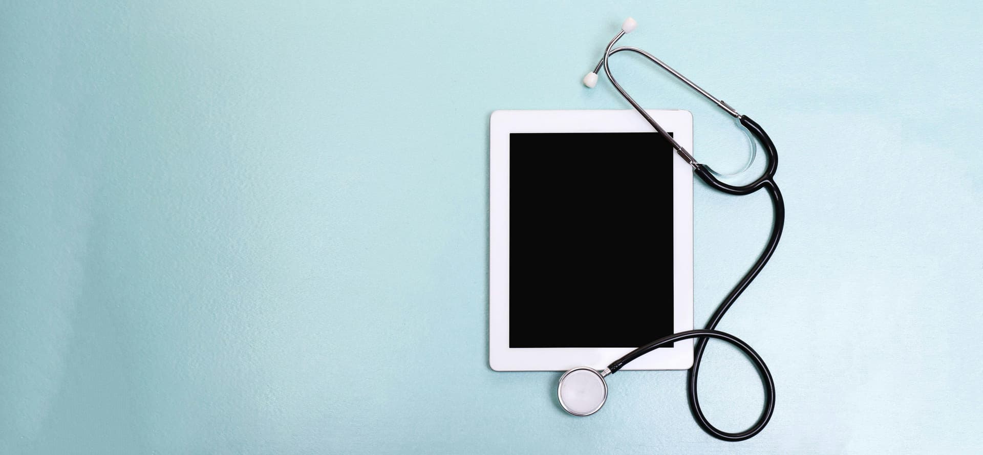 A stethoscope rests atop a tablet with a black screen, lying on a smooth, light blue surface.