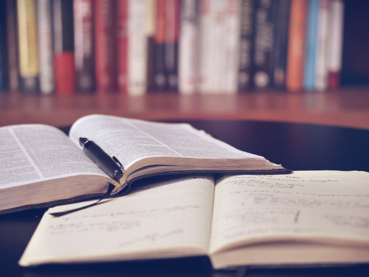 An open book holds a pen within its pages, placed on a table besides a notebook filled with handwritten notes; in the background, a filled bookshelf.
