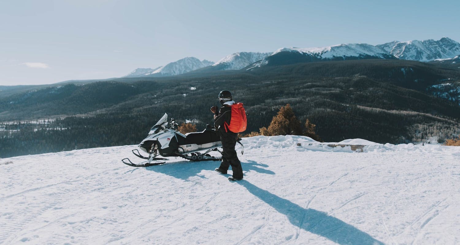 A man standing by a Skidoo, on top of a hill overlooking a valley. It’s winter.