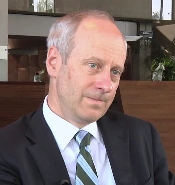 A headshot of a man in a dark suit and striped tie, sits and looks thoughtful in a modern office with wooden panels and glass windows behind him. No text present.