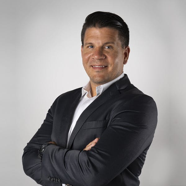 A man with short dark hair stands confidently with arms crossed, wearing a dark suit and white shirt, against a plain light gray background.