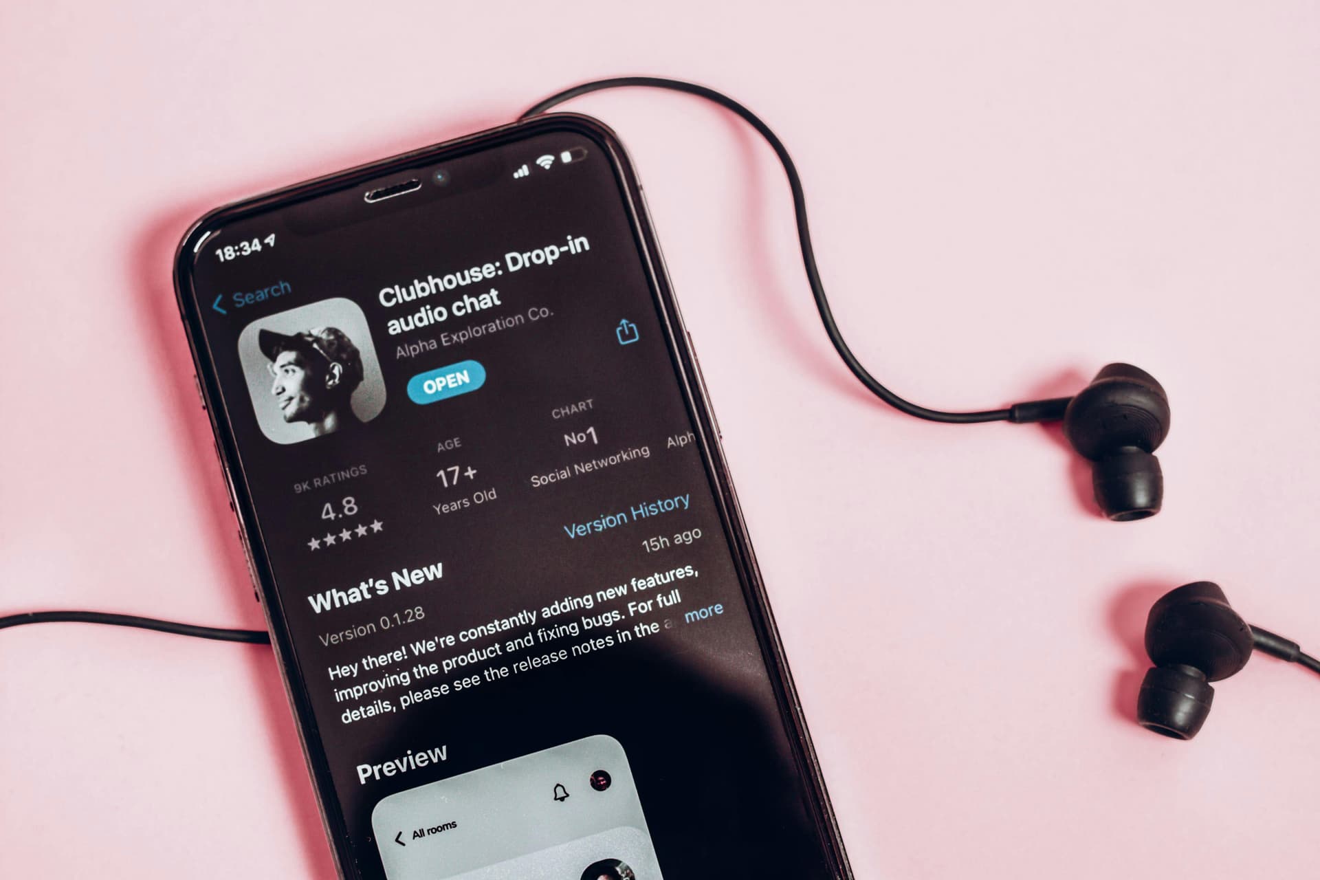 A smartphone displays the Clubhouse app page on its screen with wired black earbuds placed beside it on a pink surface. The screen shows app details and user ratings.

Text:
"Clubhouse: Drop-in audio chat
Alpha Exploration Co.
OPEN
9K RATINGS
4.8
STARS
AGE
17+
Years Old
CHART
No1
Social Networking

What’s New
Version 0.1.28
Hey there! We’re constantly adding new features, improving the product and fixing bugs. For full details, please see the release notes in the
Version History
15h ago

Preview
All rooms"
