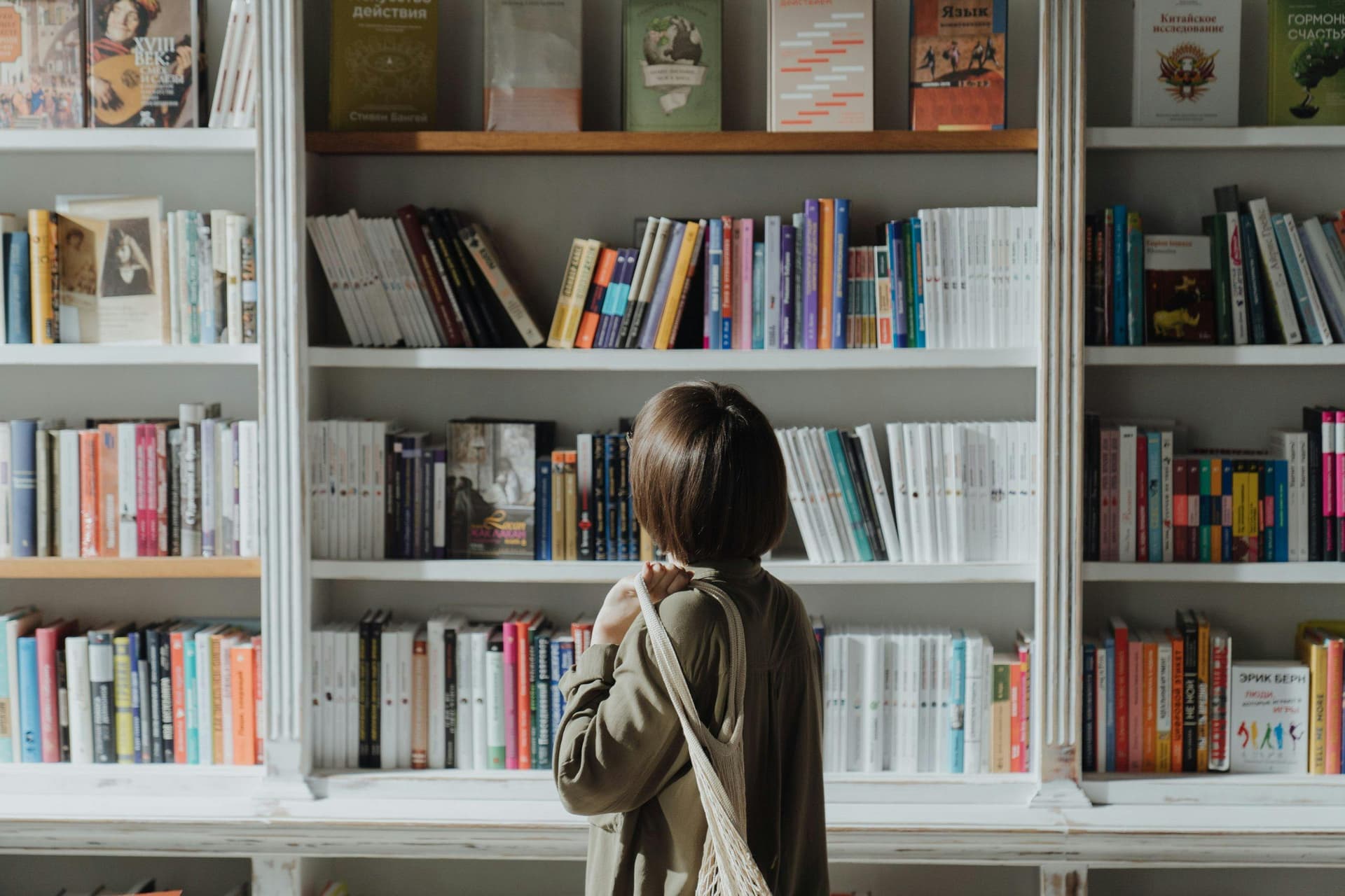 The image shows a person with short hair standing in front of a large bookshelf filled with a variety of colorful books. The individual is facing the bookshelf, holding a bag over their shoulder, seemingly browsing or contemplating which book to choose.