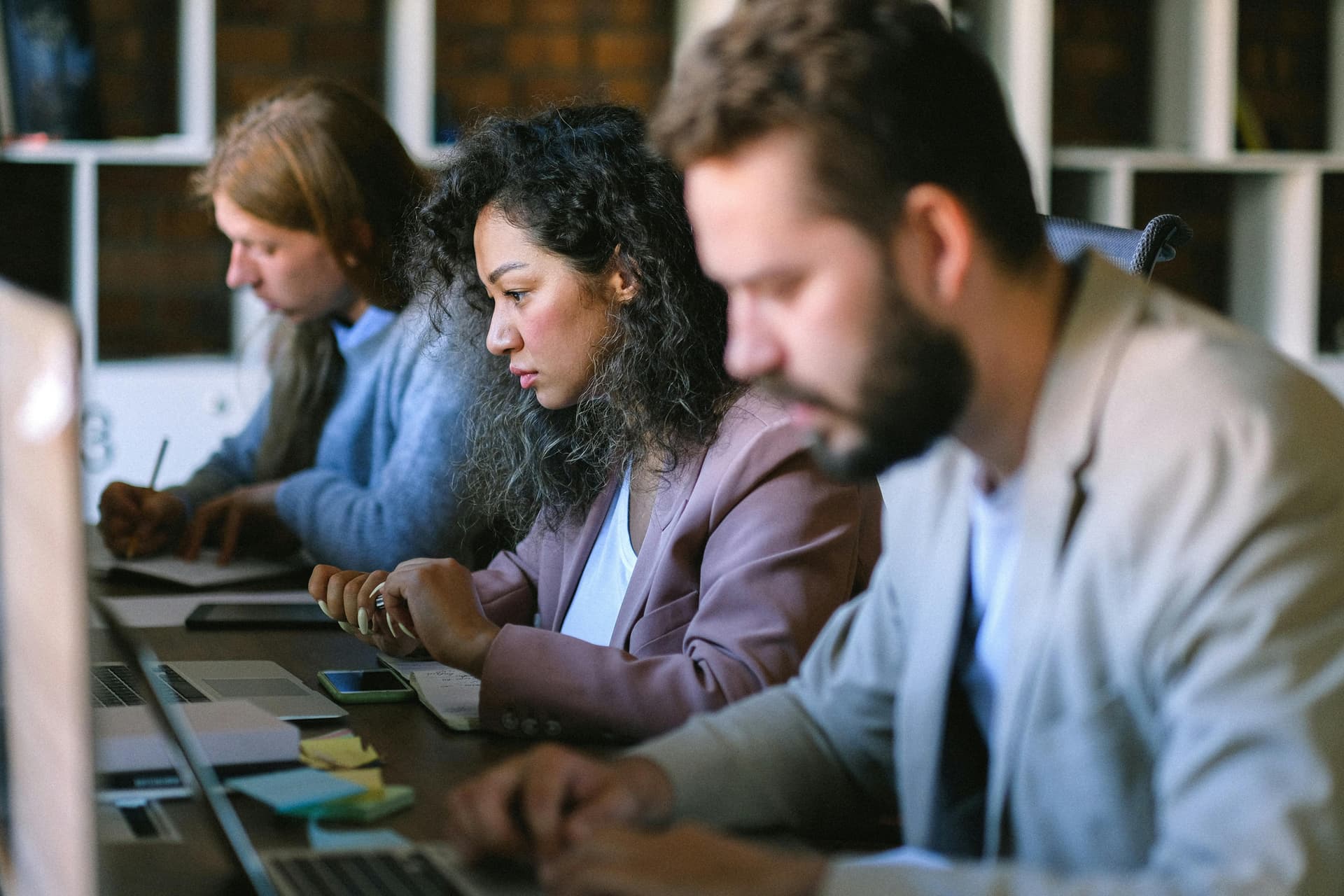 Three people are sitting at a table working on laptops and notebooks in a well-lit office with shelves in the background.