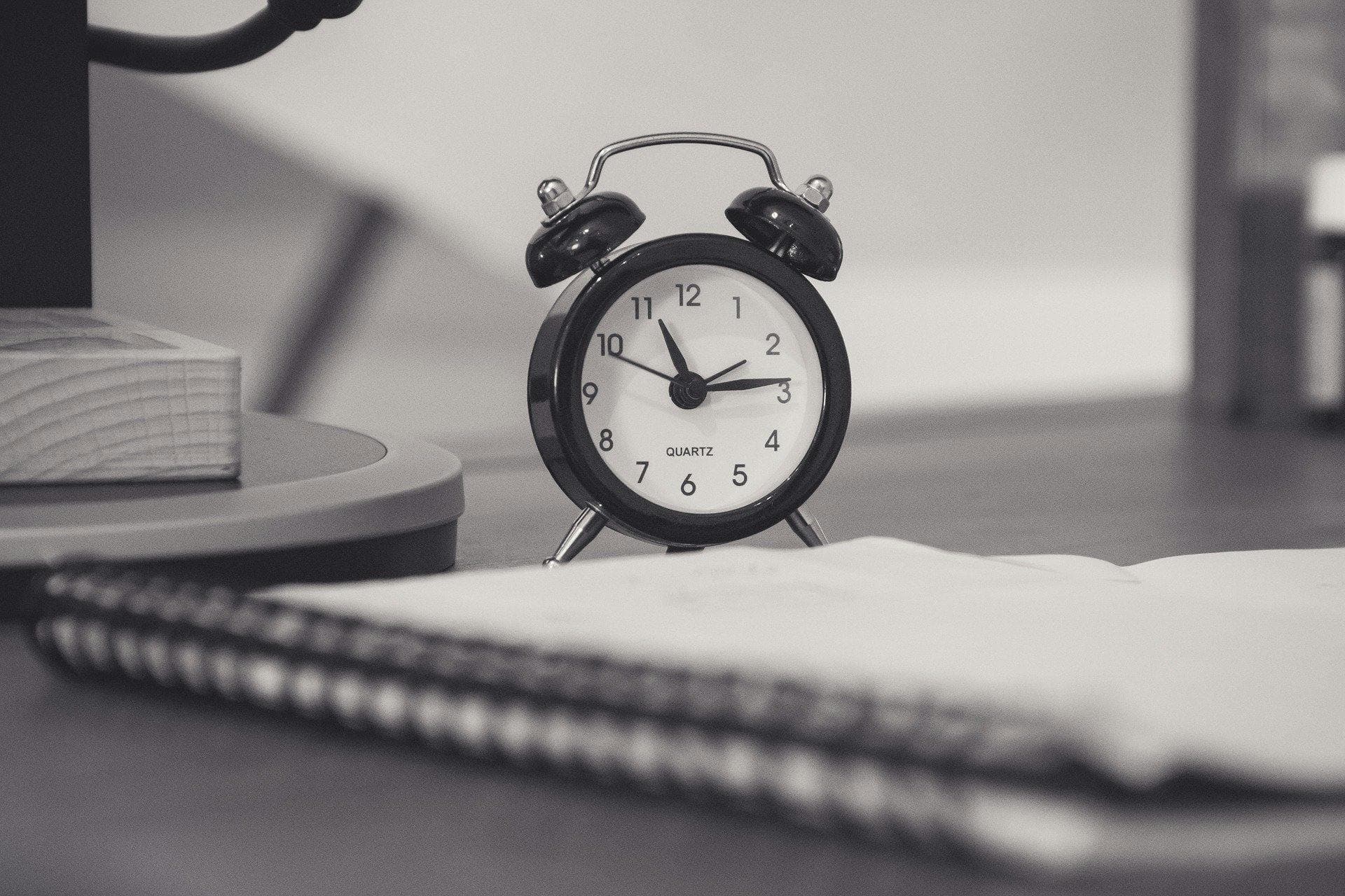 A small black alarm clock showing 10:09 stands on a wooden surface beside a spiral notebook in a dimly lit room.