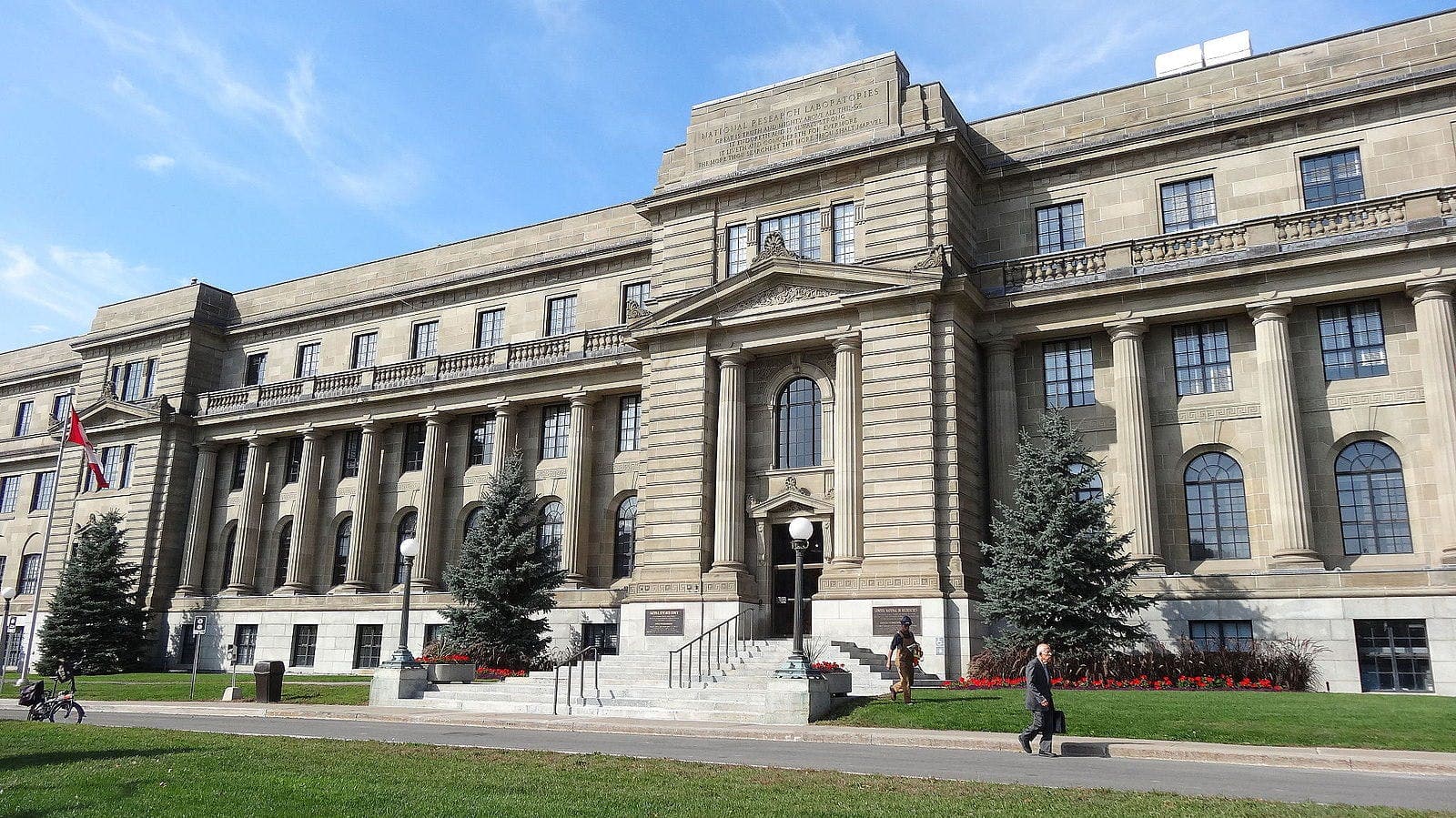 A large, historical building with columns and arched windows, titled "National Research Laboratories," stands amidst a well-maintained lawn and walkway, with people and a bicycle in the foreground.