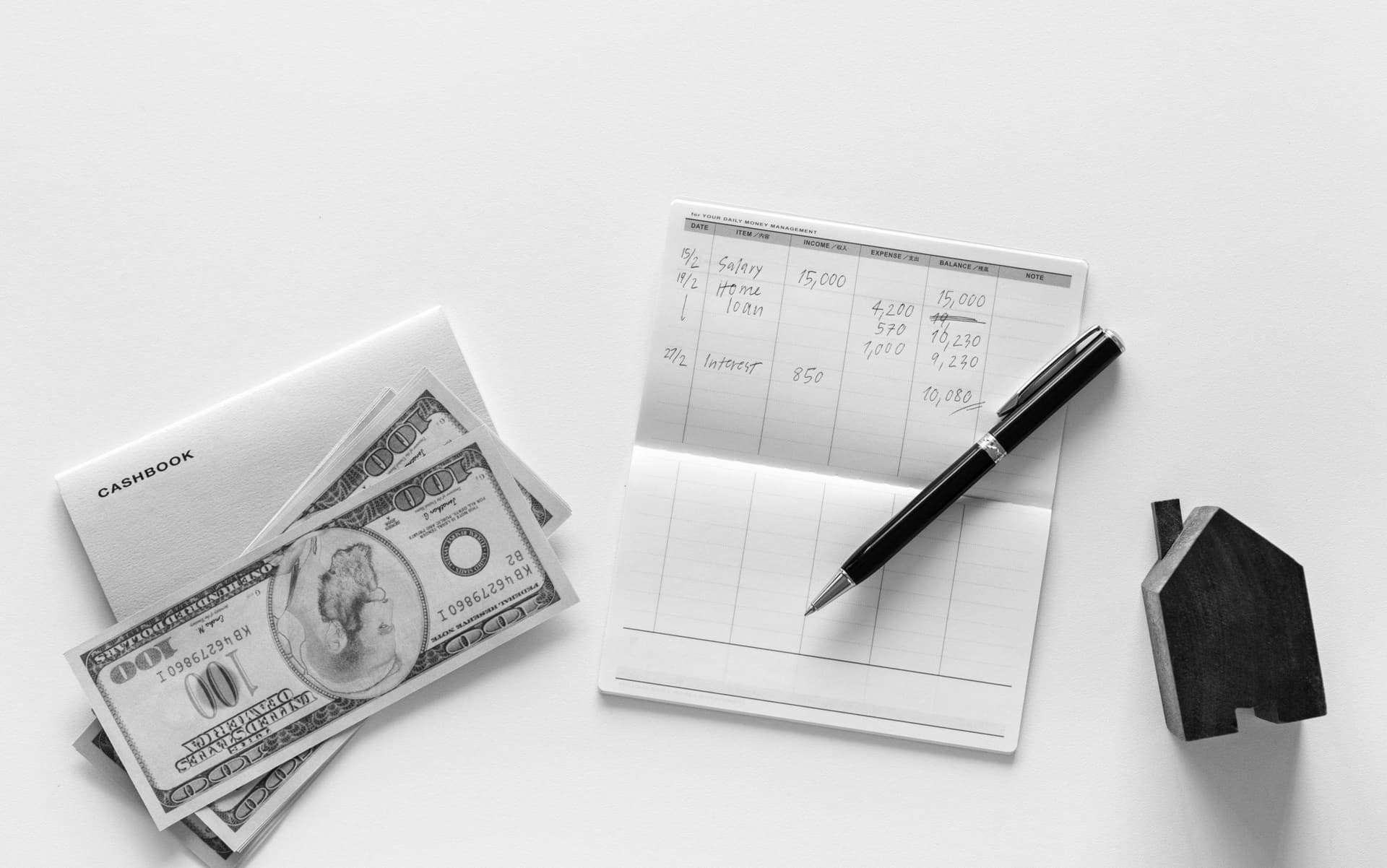 A cashbook rests with scattered 100-dollar bills on a table, while a pen lies on an open financial ledger next to a small wooden house model.