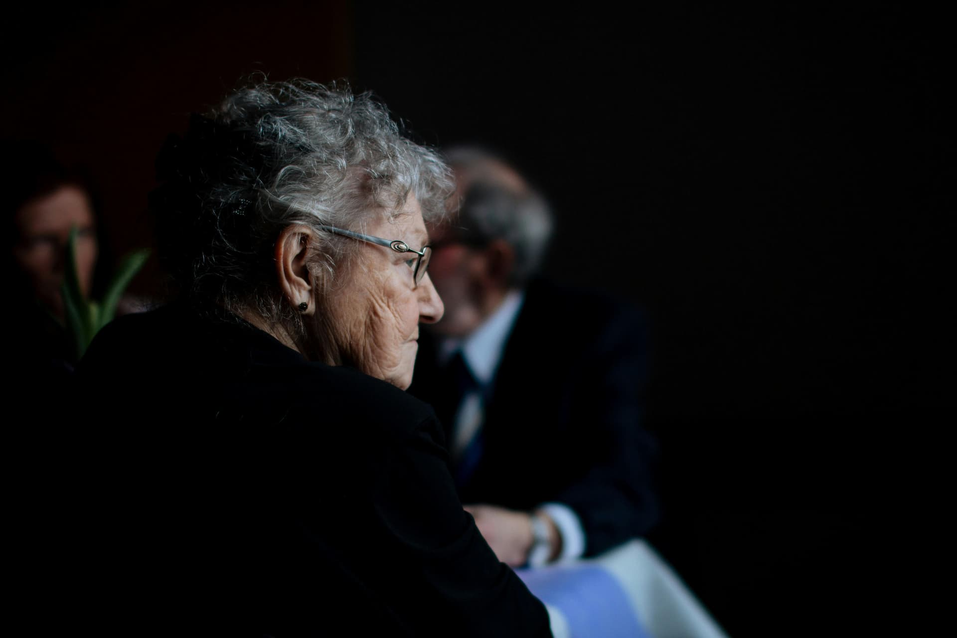 An elderly woman with gray hair and glasses sits in profile, observed in dim lighting, while another person, blurred in the background, sits at a table.