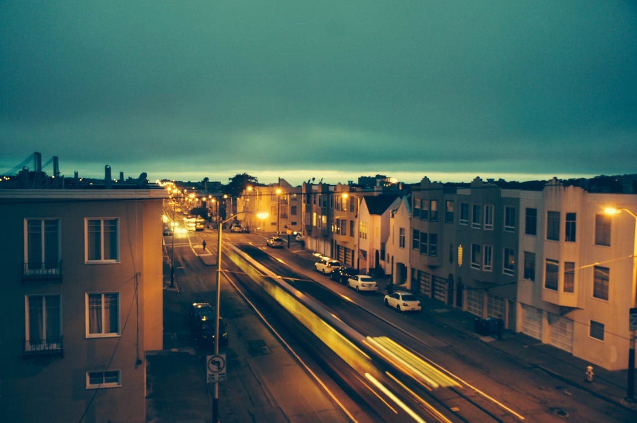 Streaks of light from moving vehicles, along a city street at dusk, with residential buildings and streetlights illuminating the surroundings.