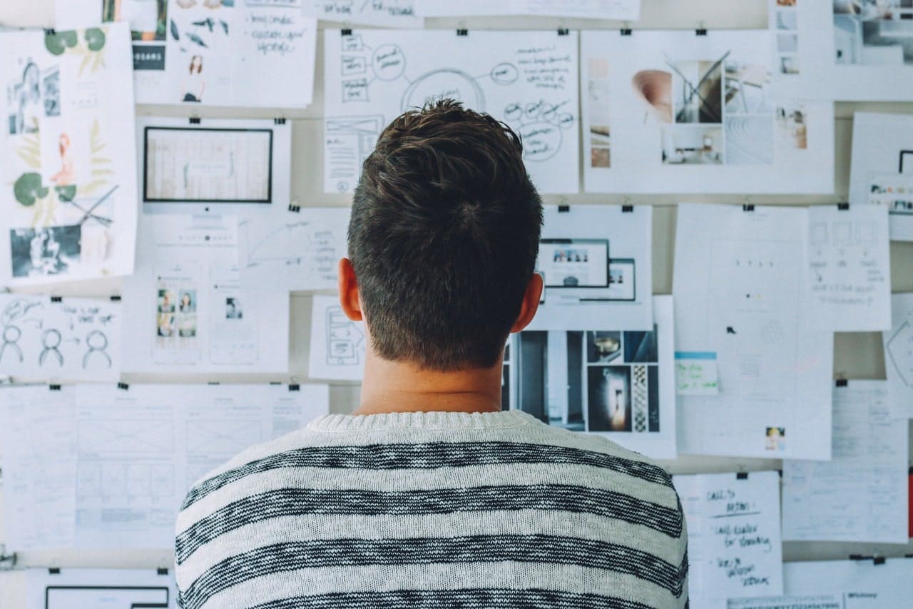 A person in a striped sweater is studying a wall covered in various papers, drawings, and notes, indicating a brainstorming or planning session.