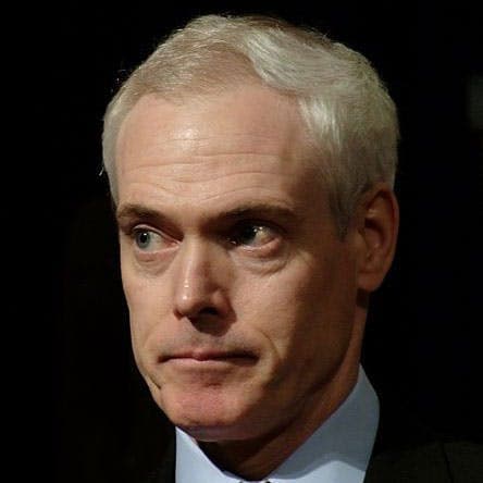 A man with short gray hair and a concerned expression looks to the side; he is wearing a dark suit and light blue shirt against a dark background.