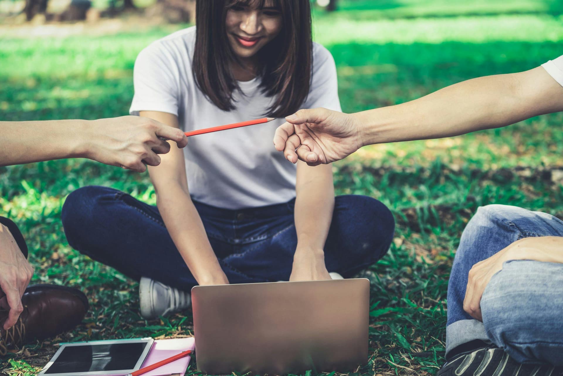 Laptop on grass with three people; two hands are passing a red pencil while a woman sits cross-legged, smiling. Nearby, a tablet and notebook lie on the ground amid a green, outdoor setting.
