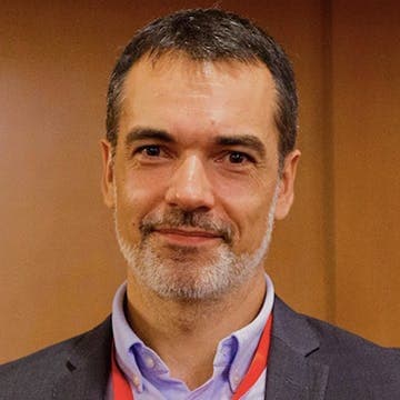 A man with short dark hair and a gray beard is smiling, wearing a suit jacket, light blue shirt, and red lanyard, standing in an indoor setting with brown walls.
