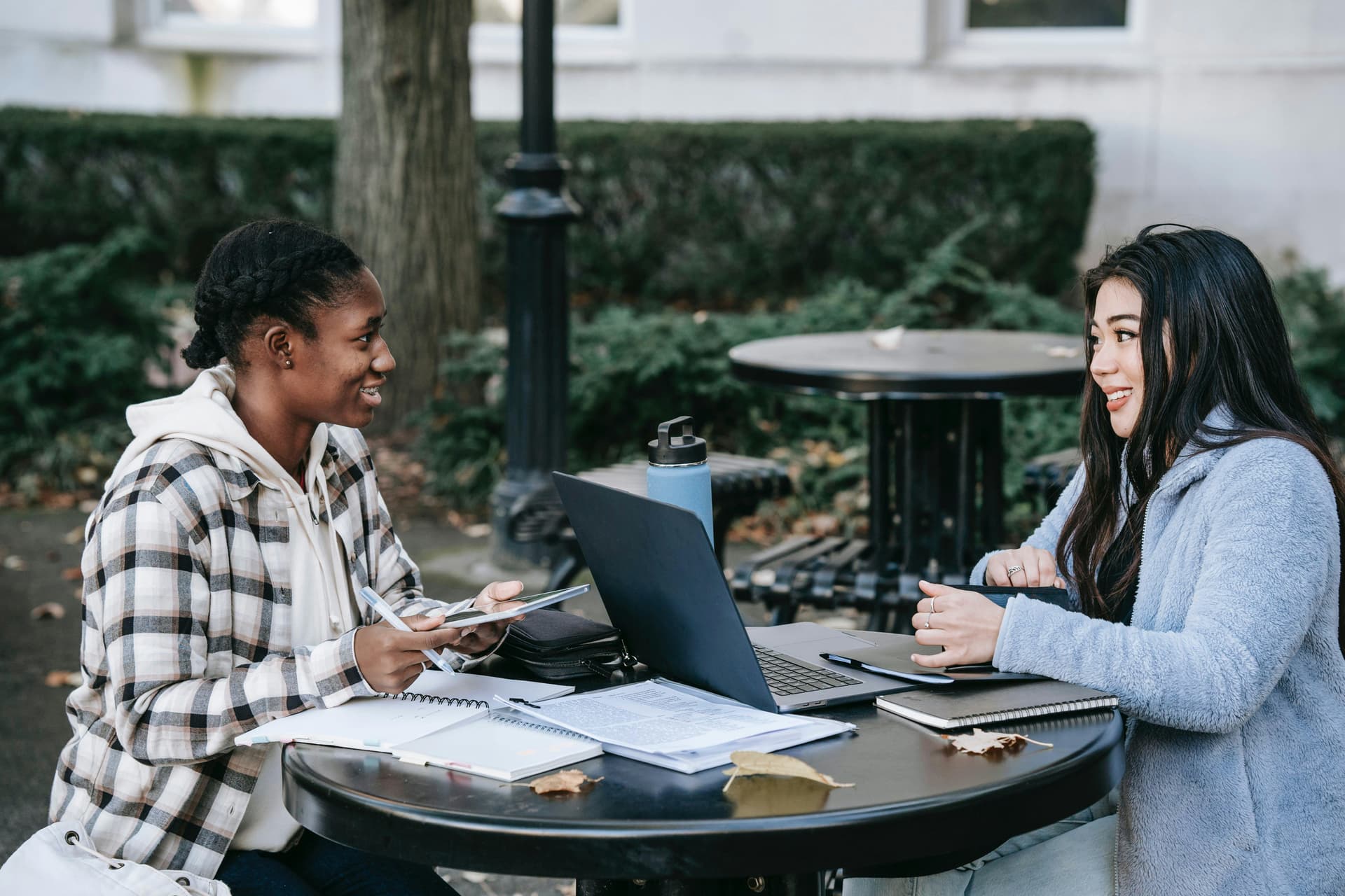 Two people converse while seated at an outdoor table with notebooks and a laptop, surrounded by greenery and a large tree.