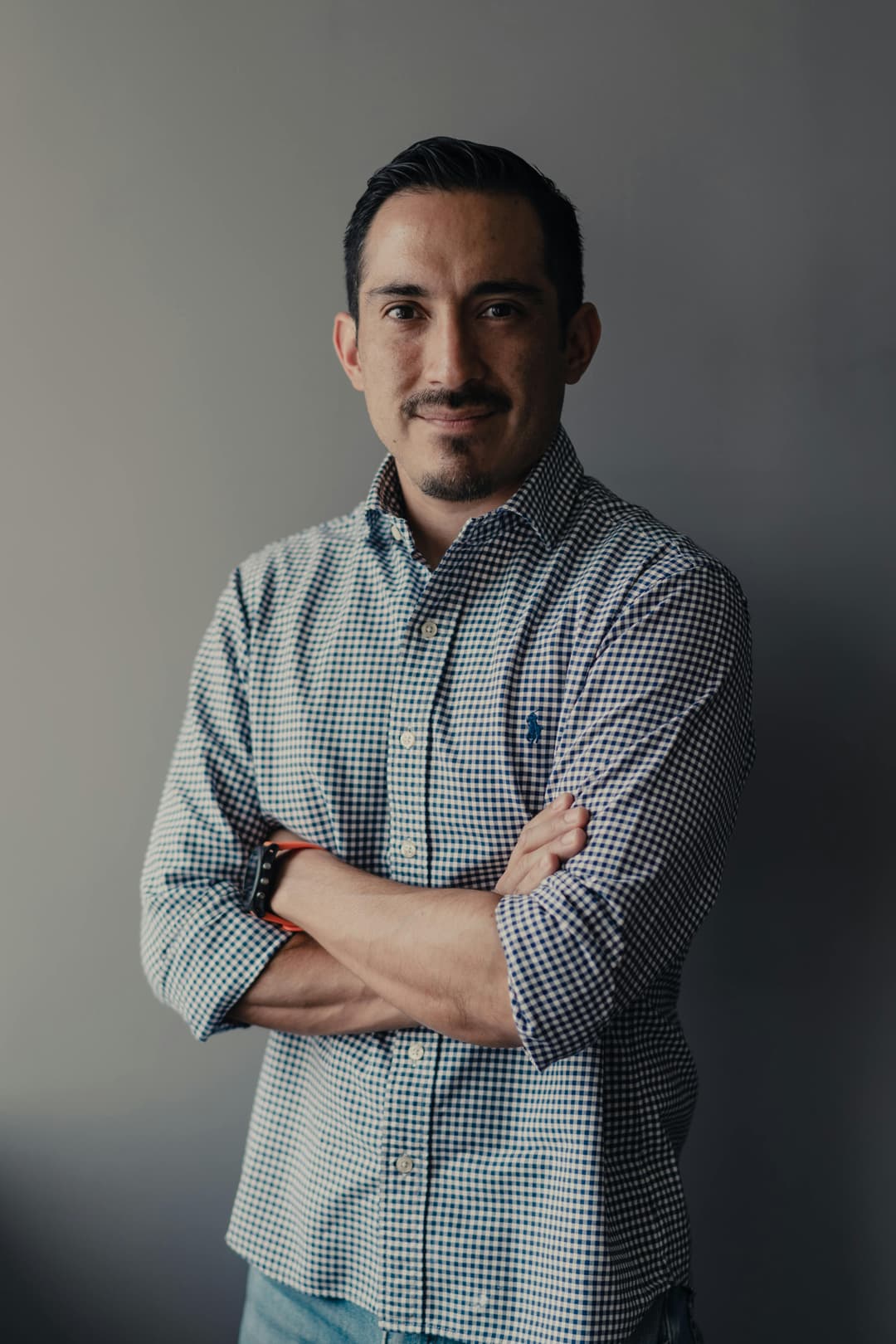 Man with short dark hair and beard standing with arms crossed, wearing a plaid dress shirt against a plain, gray background.