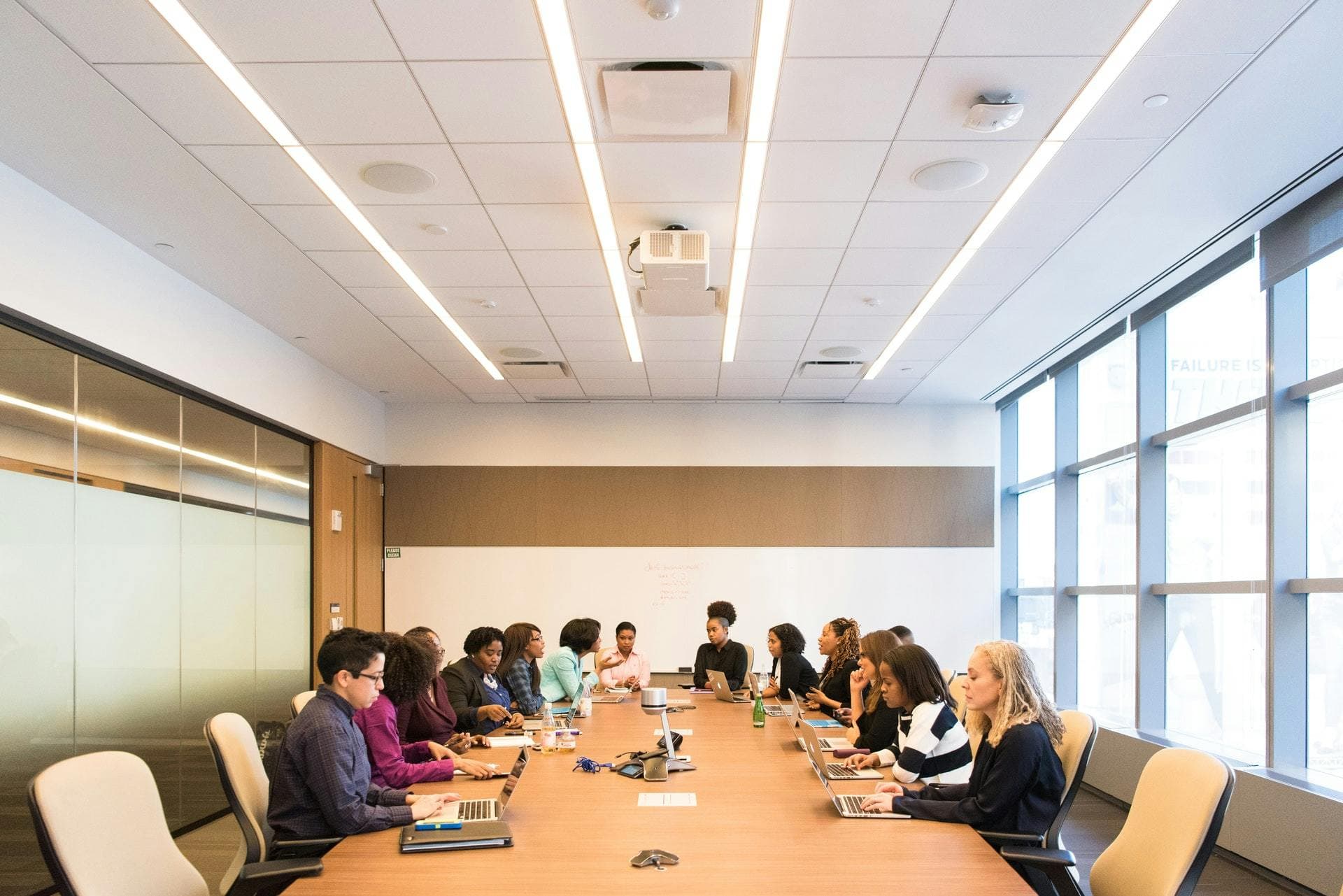 Several people sit around a large conference table using laptops in a modern office with a whiteboard and large windows. Text on whiteboard: "Created by Cody Booth."