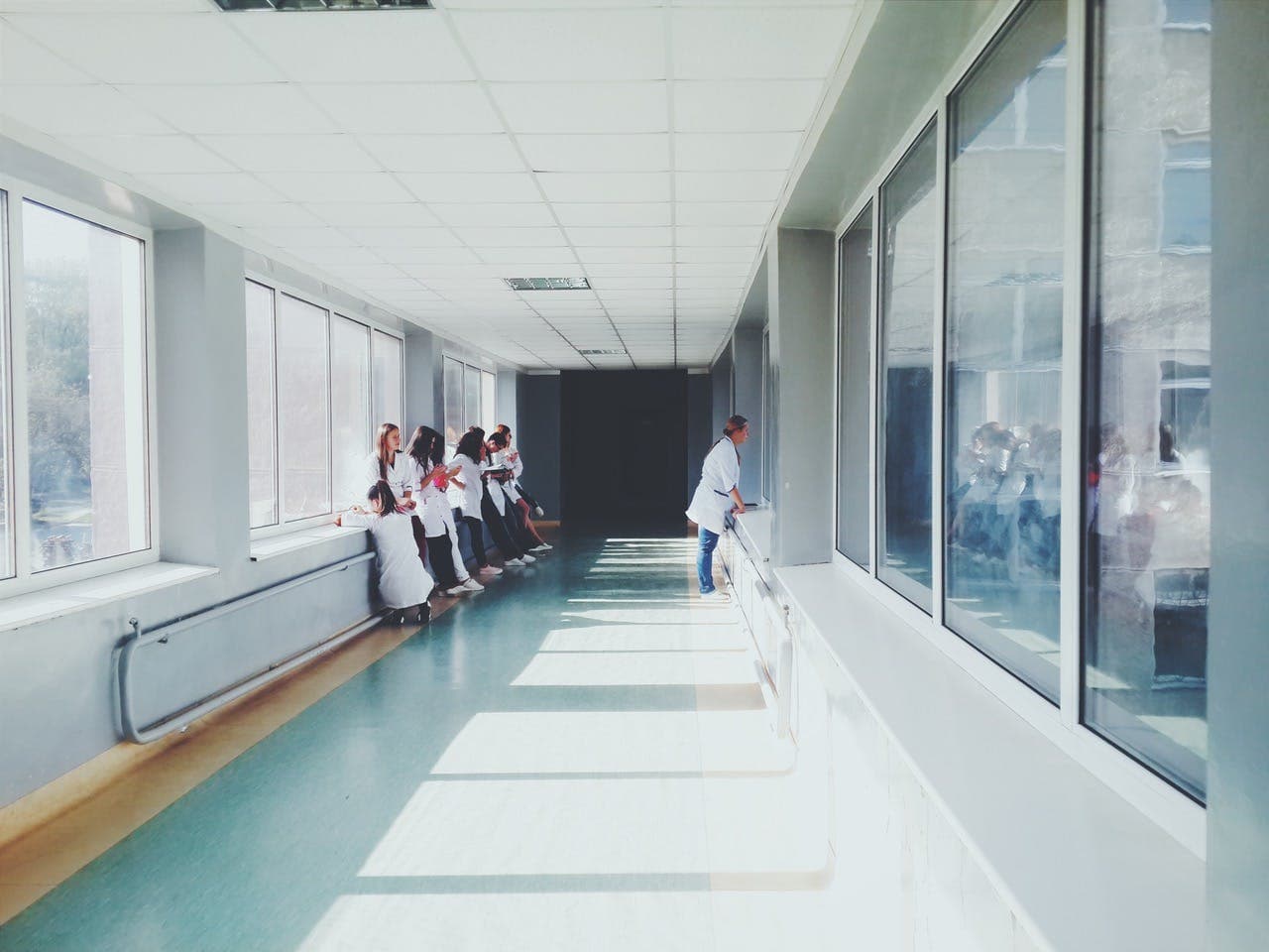 Group of people in white coats standing in a brightly lit hallway with large windows, while one person leans against the windows on the opposite side.