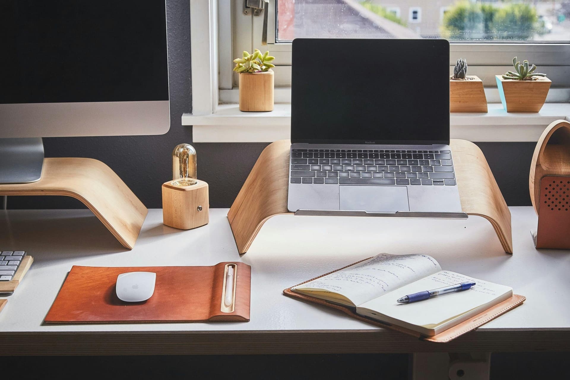 A laptop sits on a wooden stand on a desk with an open notebook and pen, a mouse on a leather pad, monitor, and small plants near a window.