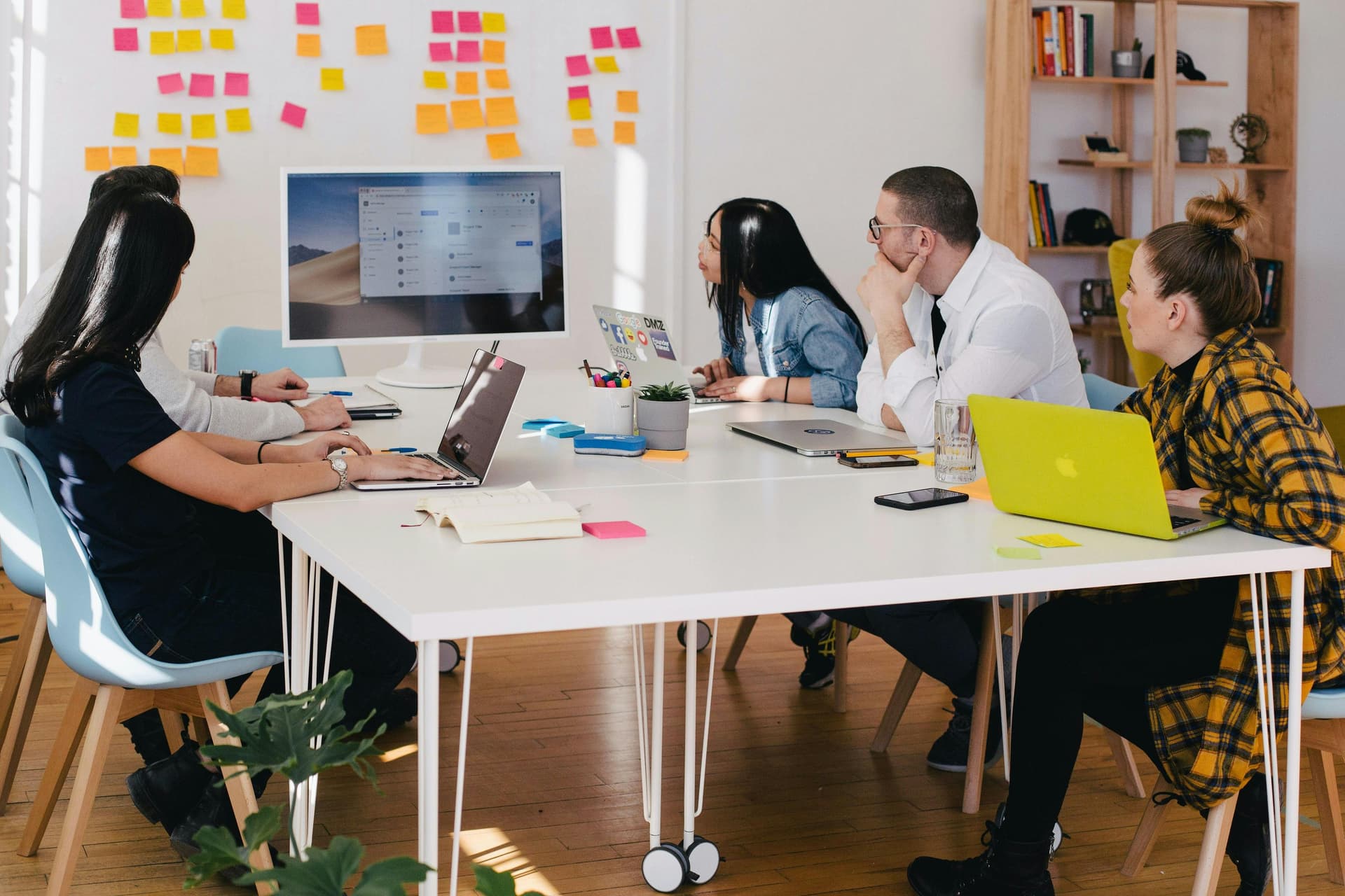 Team in a meeting room, with a woman standing and pointing at sticky notes on a whiteboard, while others sit around a table with laptops.