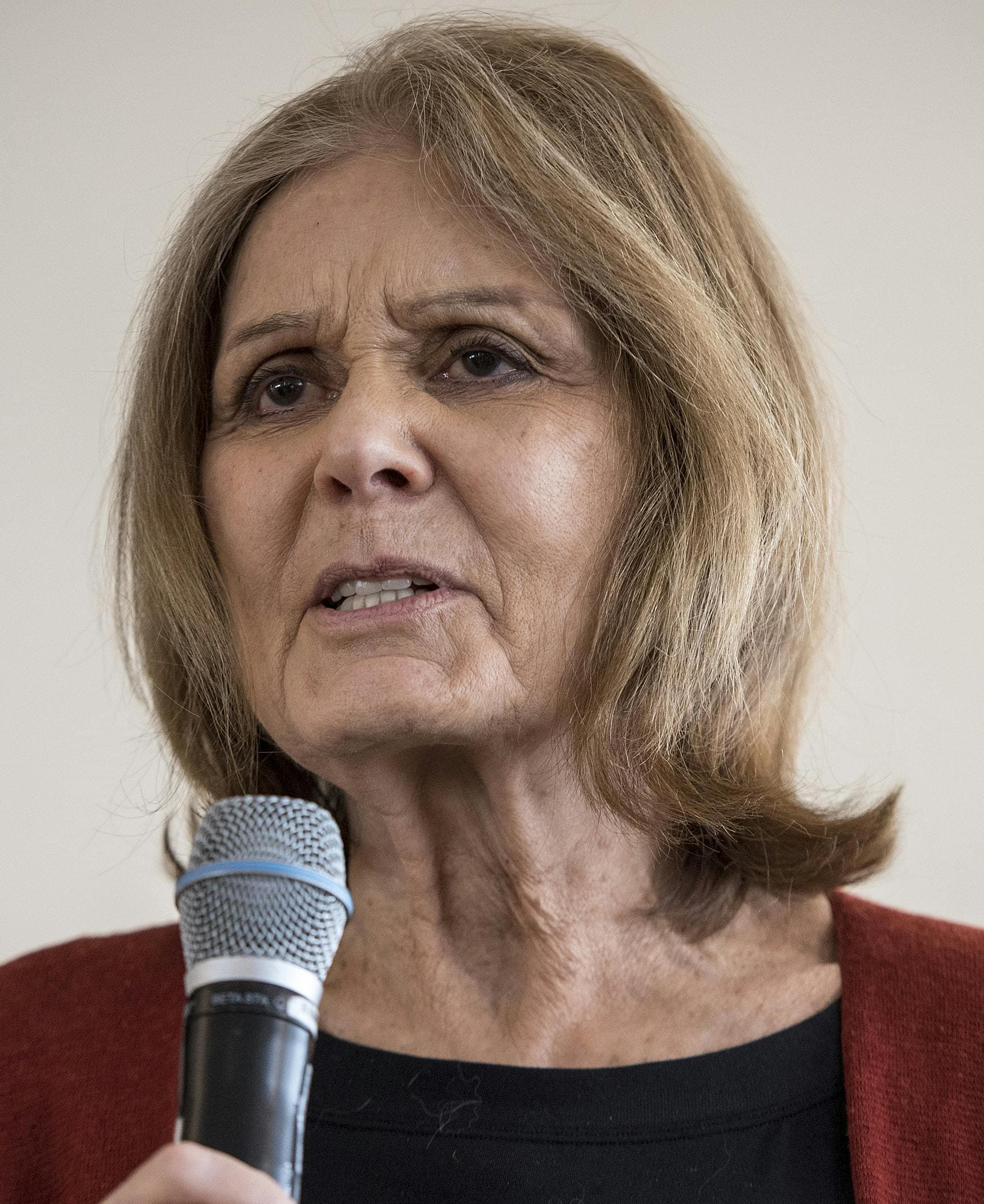 Headshot of a woman with blond hair, holding a microphone. She is wearing a black top and a red cardigan.