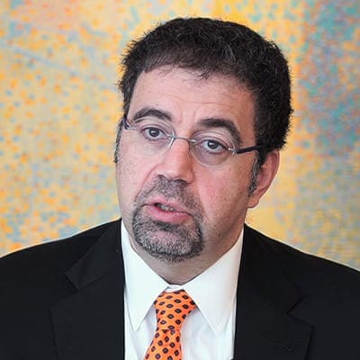 A man in a black suit and orange tie speaks against a colorful, abstract background. He has glasses and short, dark hair. 

[No text found]