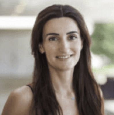 A woman with long dark hair smiles softly, standing outdoors with a blurred background of green foliage and light-colored structures.