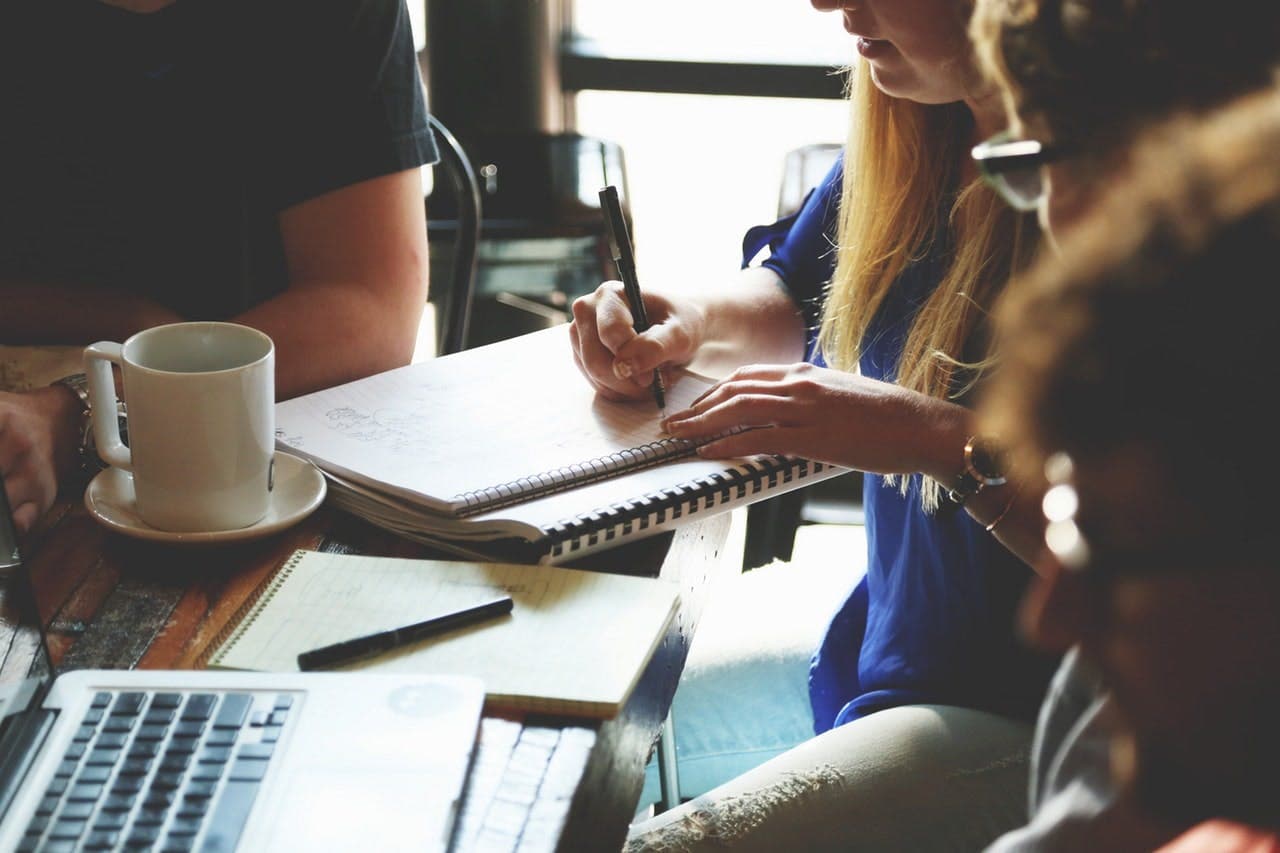 People seated around a table, writing in notebooks and using a laptop, with coffee cups nearby, suggesting a casual meeting or collaborative work session.