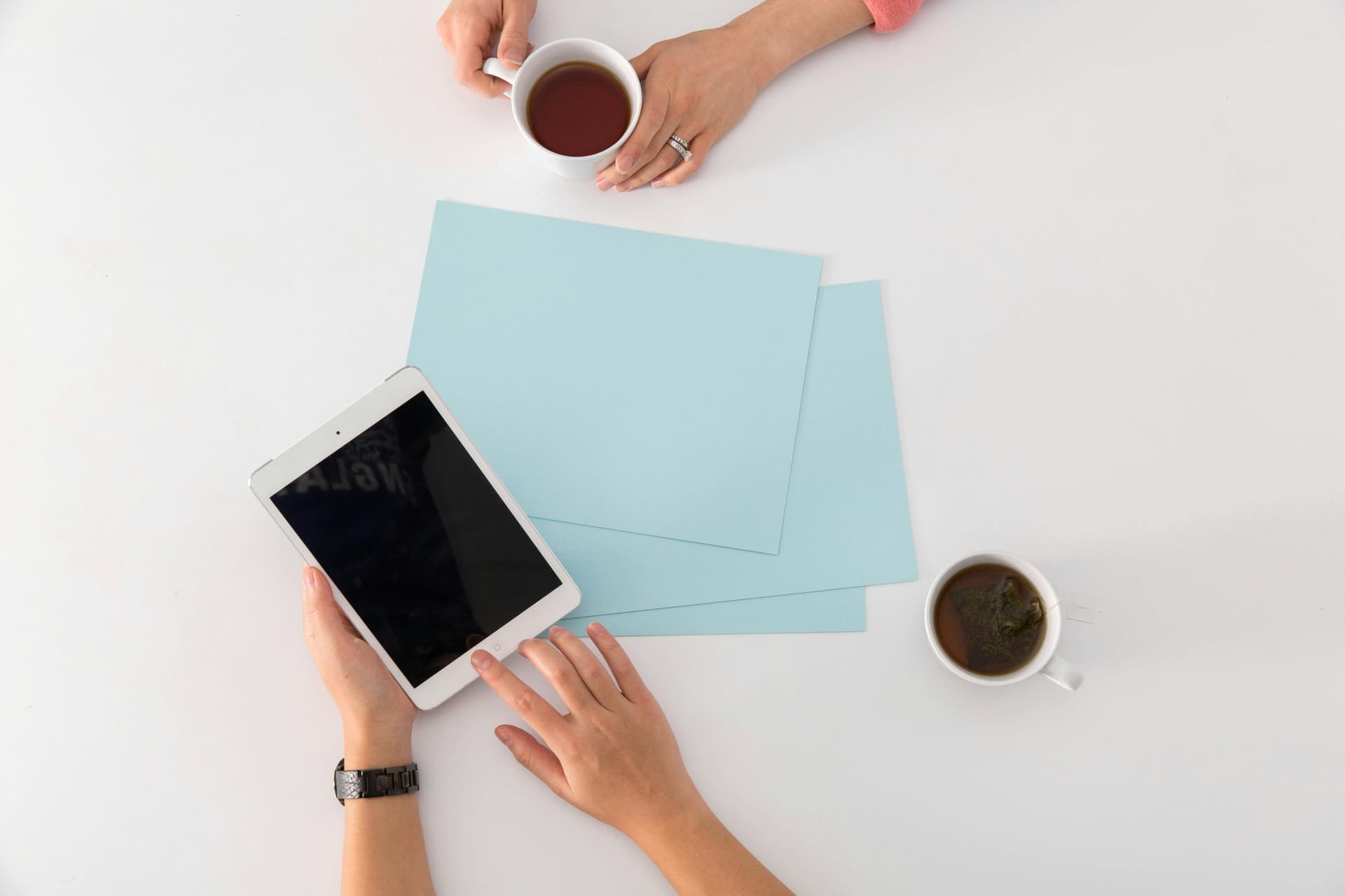 Two hands holding cups of tea and one hand holding a tablet, around blue documents on a white table.