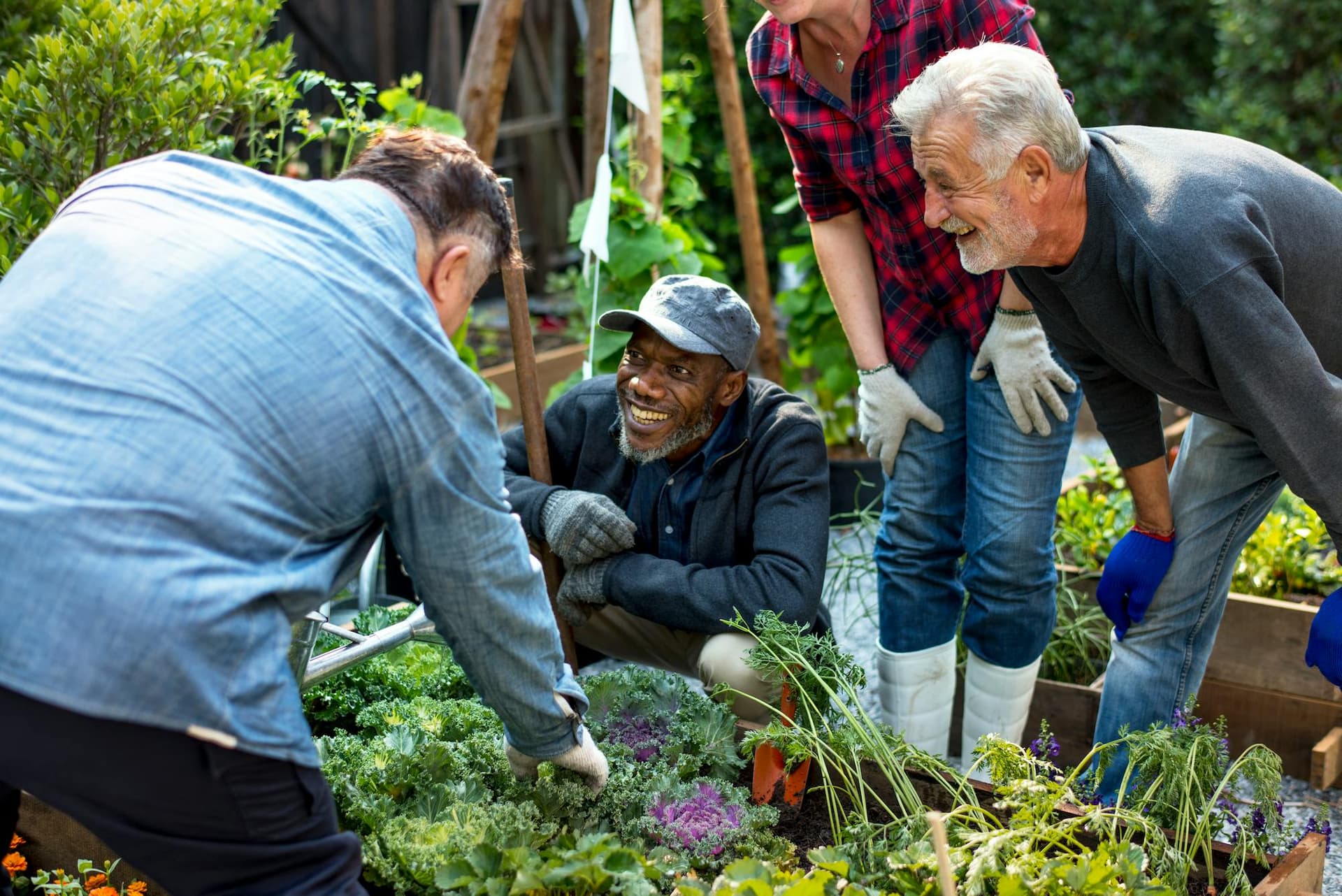 a group of four people gardening together. They appear to be of varying ages, smiling and engaging with each other. One man is crouched down while others stand nearby, and they are surrounded by greenery and plants in what looks like a community garden