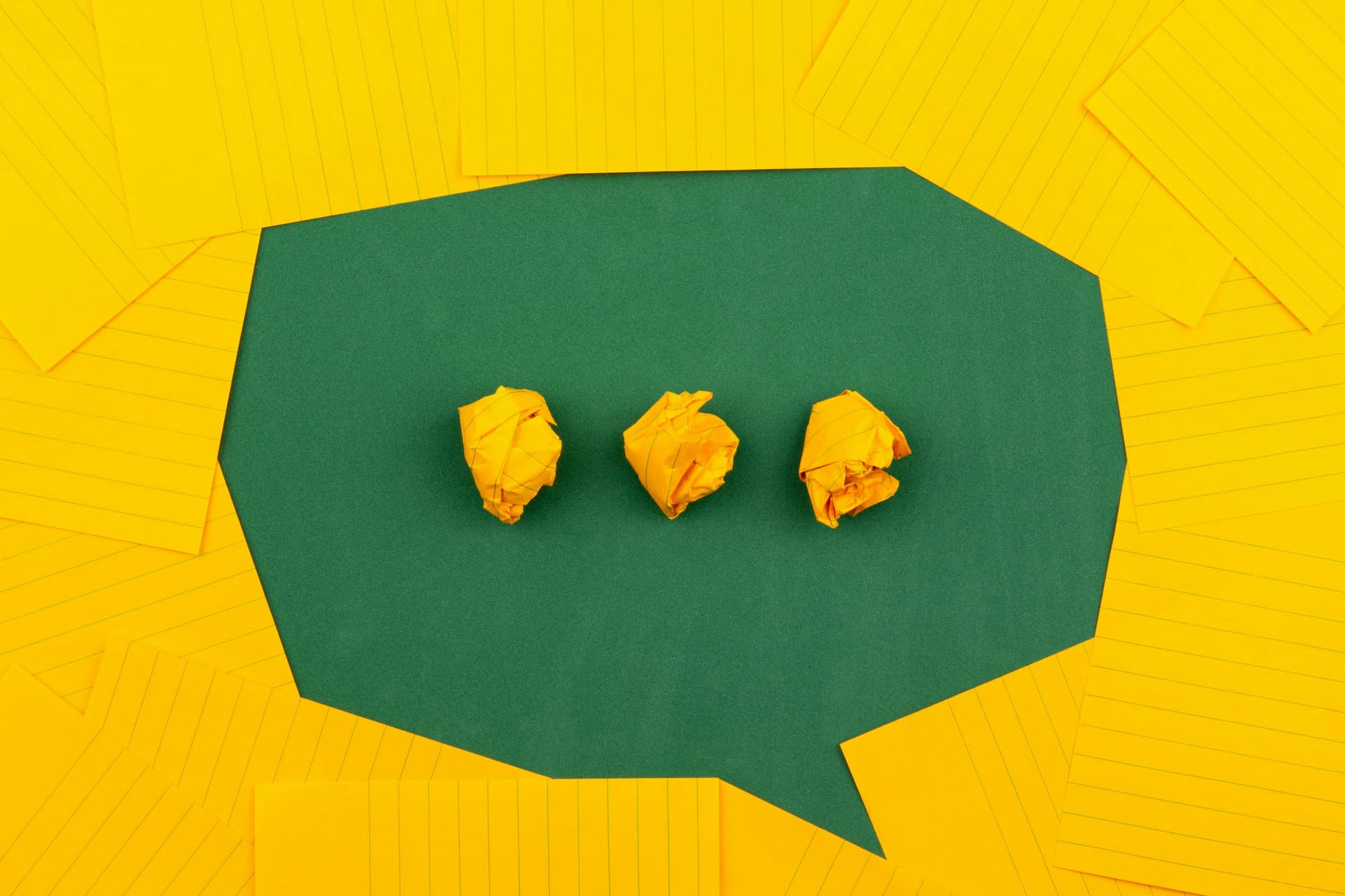 Yellow crumpled paper balls arranged in a row on a green background shaped like a speech bubble, surrounded by neatly placed yellow lined papers.