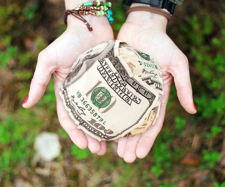 Hands cradle a crumpled ball made of $100 bills adorned with bracelets in an outdoor setting with green vegetation.