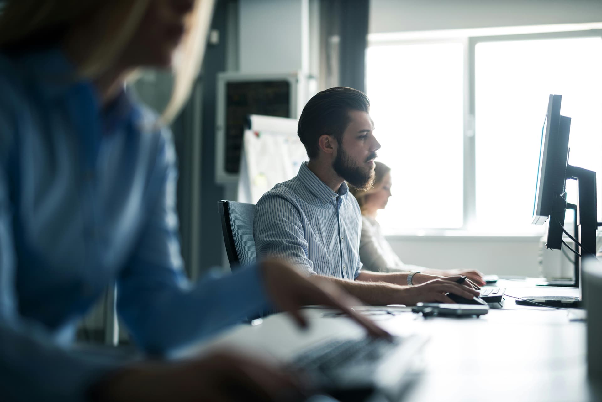 A photograph of three people working at computers in an office setting. The focus is on a man with a beard and short hair in the middle of the image, who is concentrating on his screen. In the foreground, a blurred figure is seen working on a laptop, while in the background, another person is also focused on their computer. The setting is well-lit with natural light coming from a large window.
