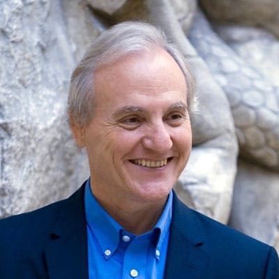 Smiling man wearing a blue shirt and dark blazer, standing against a textured stone background with hexagonal patterns.