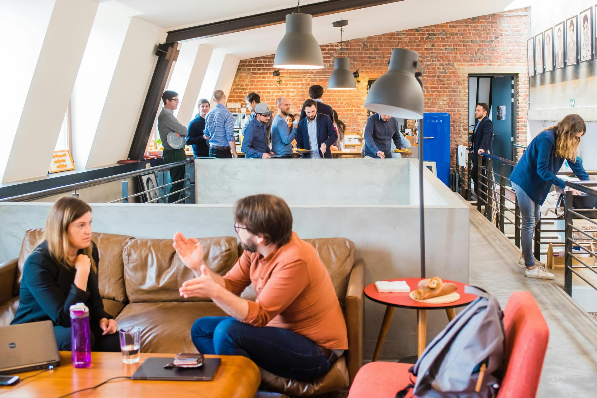 People conversing and eating in a modern office lounge with a brick wall and industrial decor; two individuals sit on a couch in the foreground while a group stands at a kitchen counter in the background.