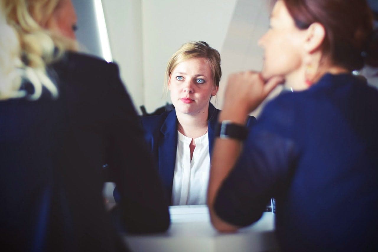 Person attentively listening to two individuals during a meeting in a well-lit room, with a white wall and minimalistic decor in the background.