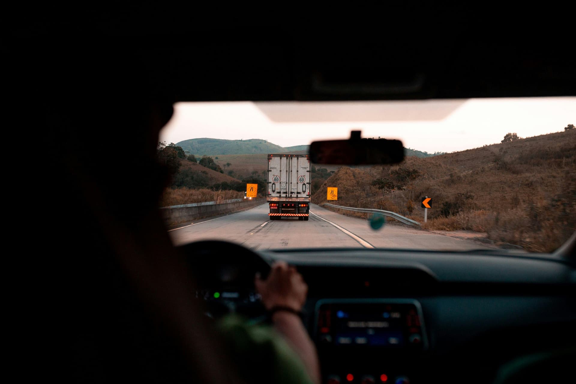 A person in a car drives on a highway behind a truck, with hills and road signs visible in the background.