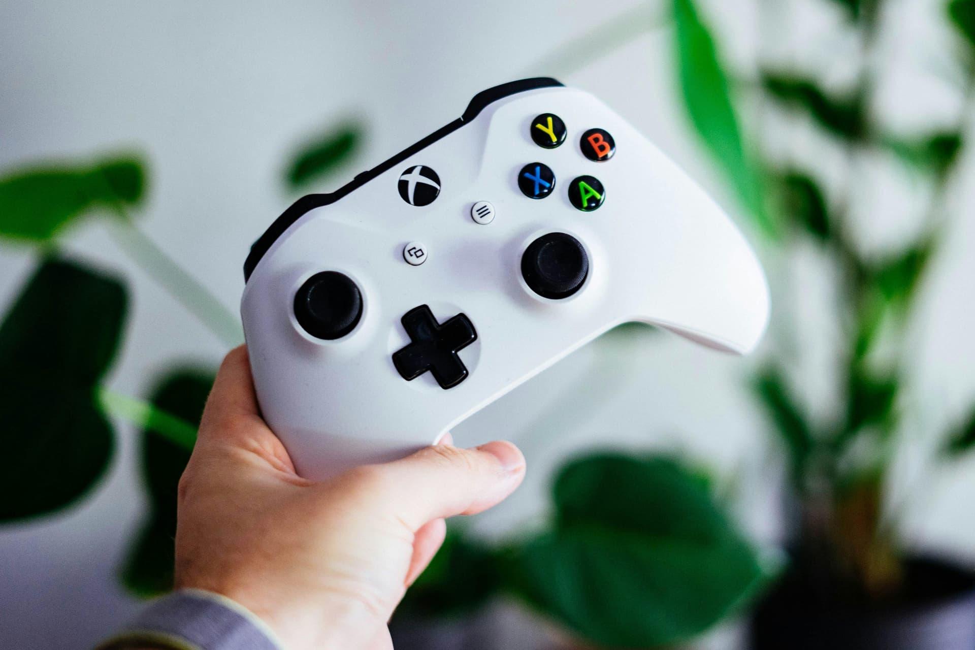 A close-up of a hand holding a white Xbox controller, with the buttons X, Y, A, and B visible in different colors. The background is blurred with green plants in soft focus.
