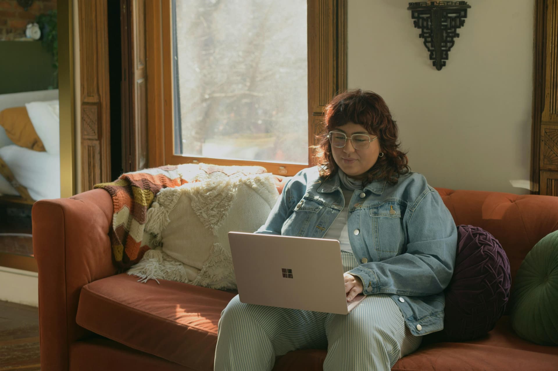 A person is sitting on an orange couch, using a laptop, next to a window with sunlight streaming in, and surrounded by cushions and cozy decor.