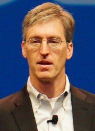 Man with short grayish hair, wearing glasses, a dark suit jacket, and a collared shirt, speaking into a microphone while looking ahead.