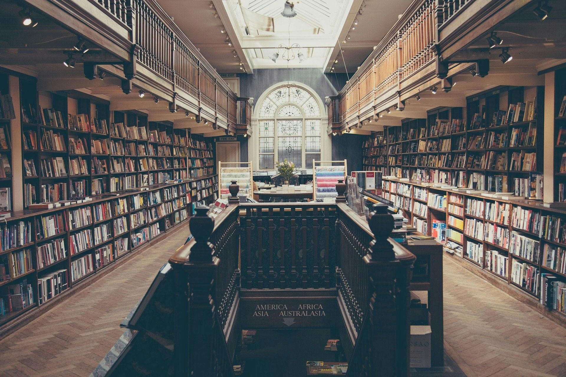 Books fill wooden shelves in a spacious library with a skylight. At the center, a wooden railing surrounds a staircase. Signs include: "AMERICA," "ASIA," "AFRICA," "AUSTRALASIA."