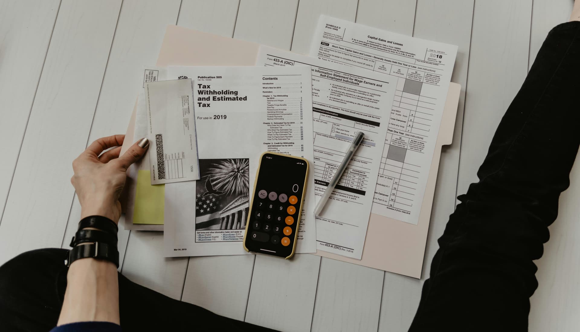A person holding a calculator, surrounded by tax forms and documents on a light-colored wooden floor. Text includes "Tax Withholding and Estimated Tax For use in 2019."