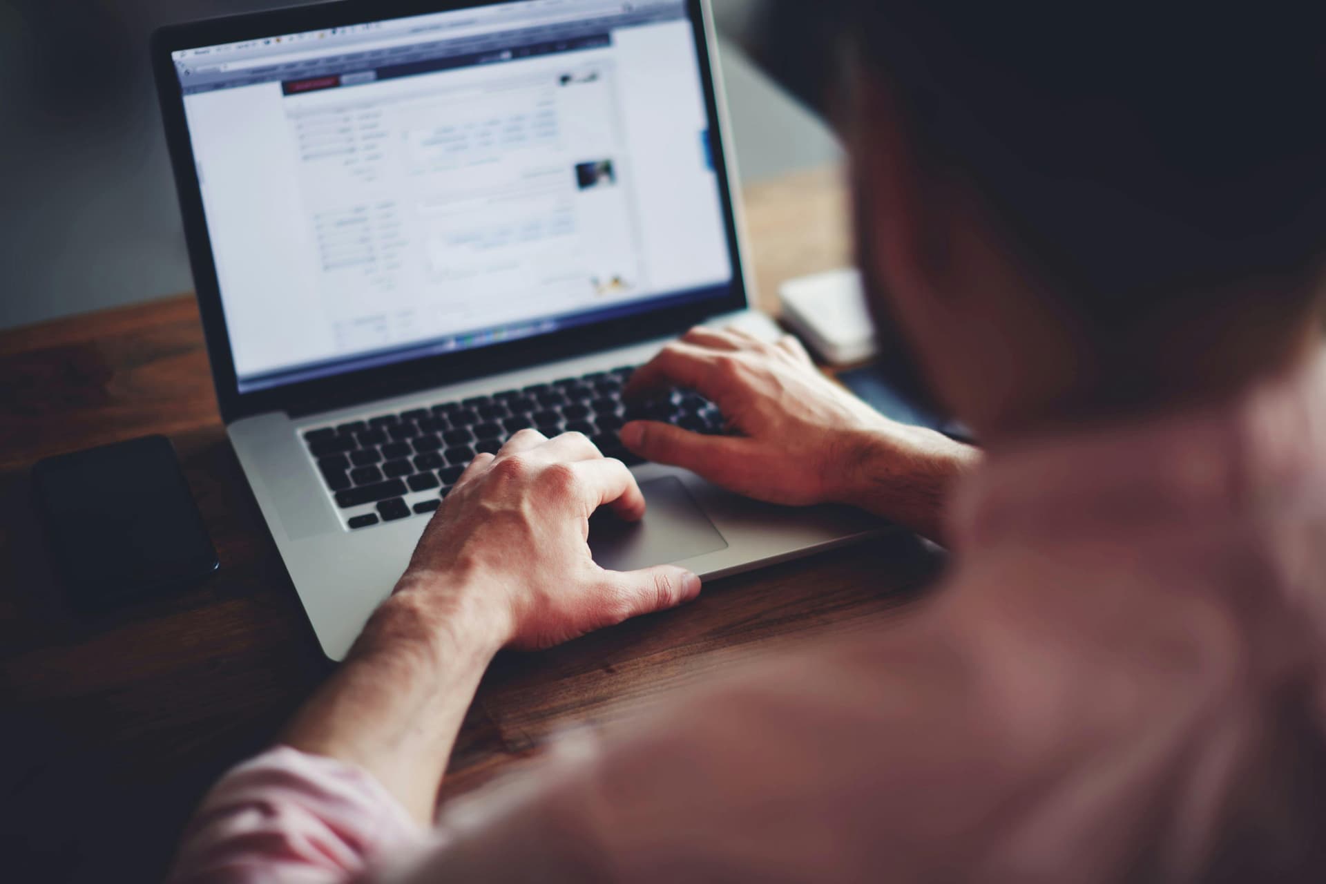 A person types on a laptop displaying a webpage with text, sitting at a wooden desk with a smartphone nearby.