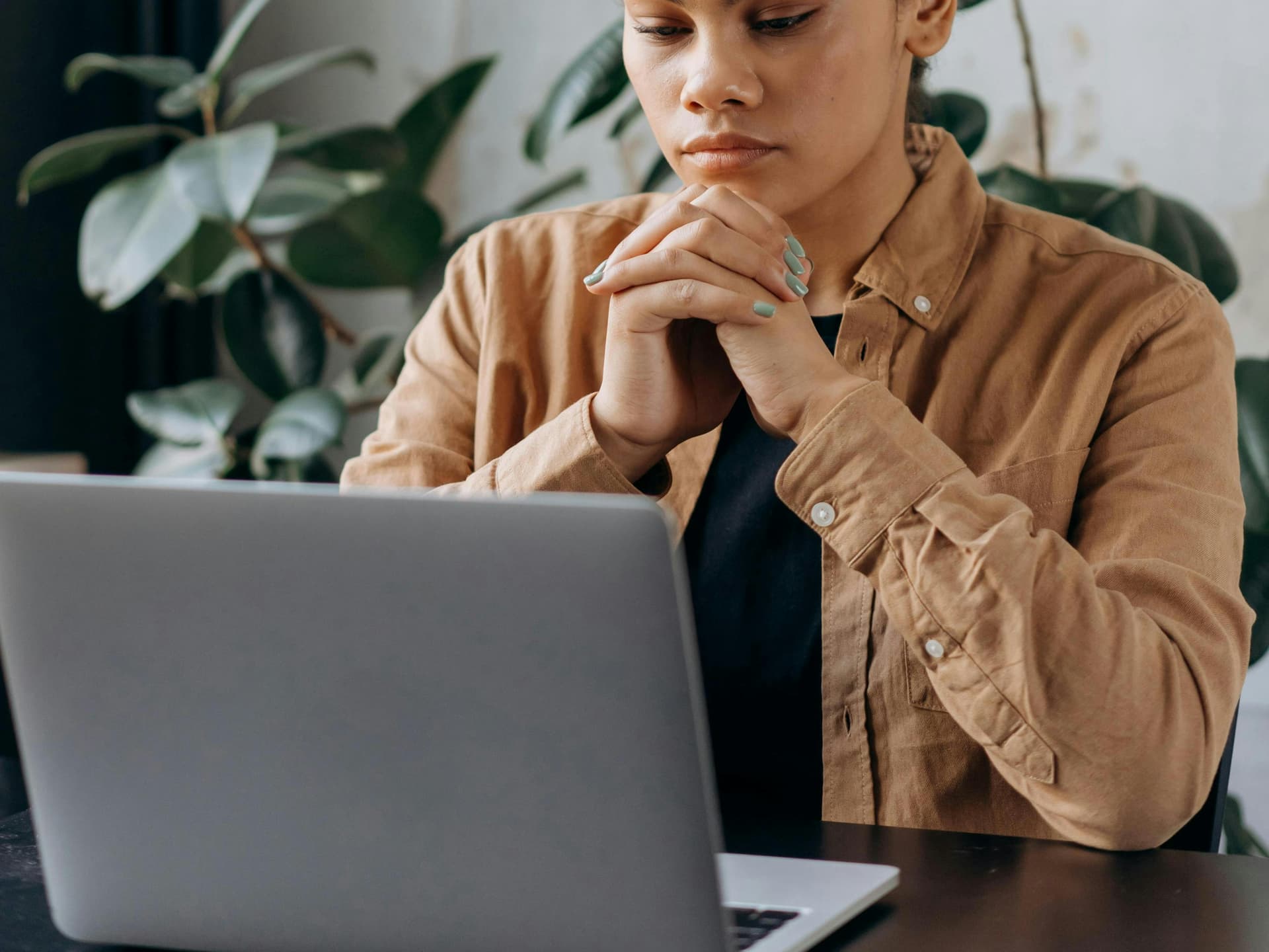 A woman of color wearing a brown vest looking at a laptop with her hands together under her chin.