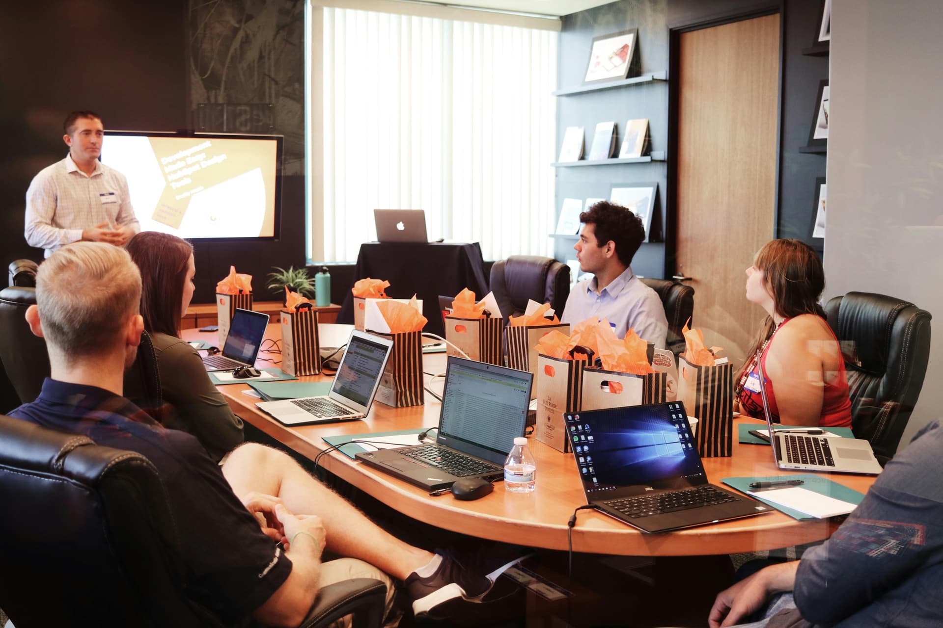 A man presents at a conference table with laptops, gift bags, and attendees focused in a modern office with shelves and a large window.