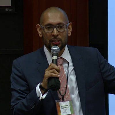 A man in a suit speaks into a microphone while standing indoors. He wears glasses, a nametag around his neck, and appears to be giving a presentation or speech.