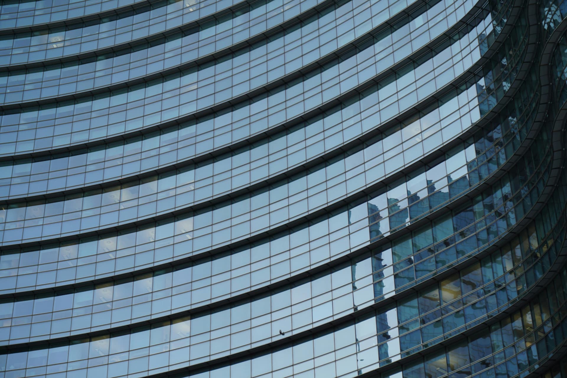 Curved glass windows reflecting the sky and surrounding skyscrapers create a sleek, modern facade on a multi-story building in an urban environment.
