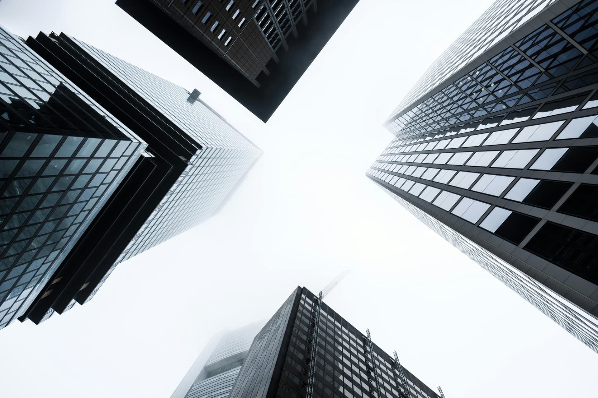 Tall skyscrapers converge upward into a cloudy, misty sky, creating a symmetrical, towering cityscape with reflective glass facades and modern architectural designs.