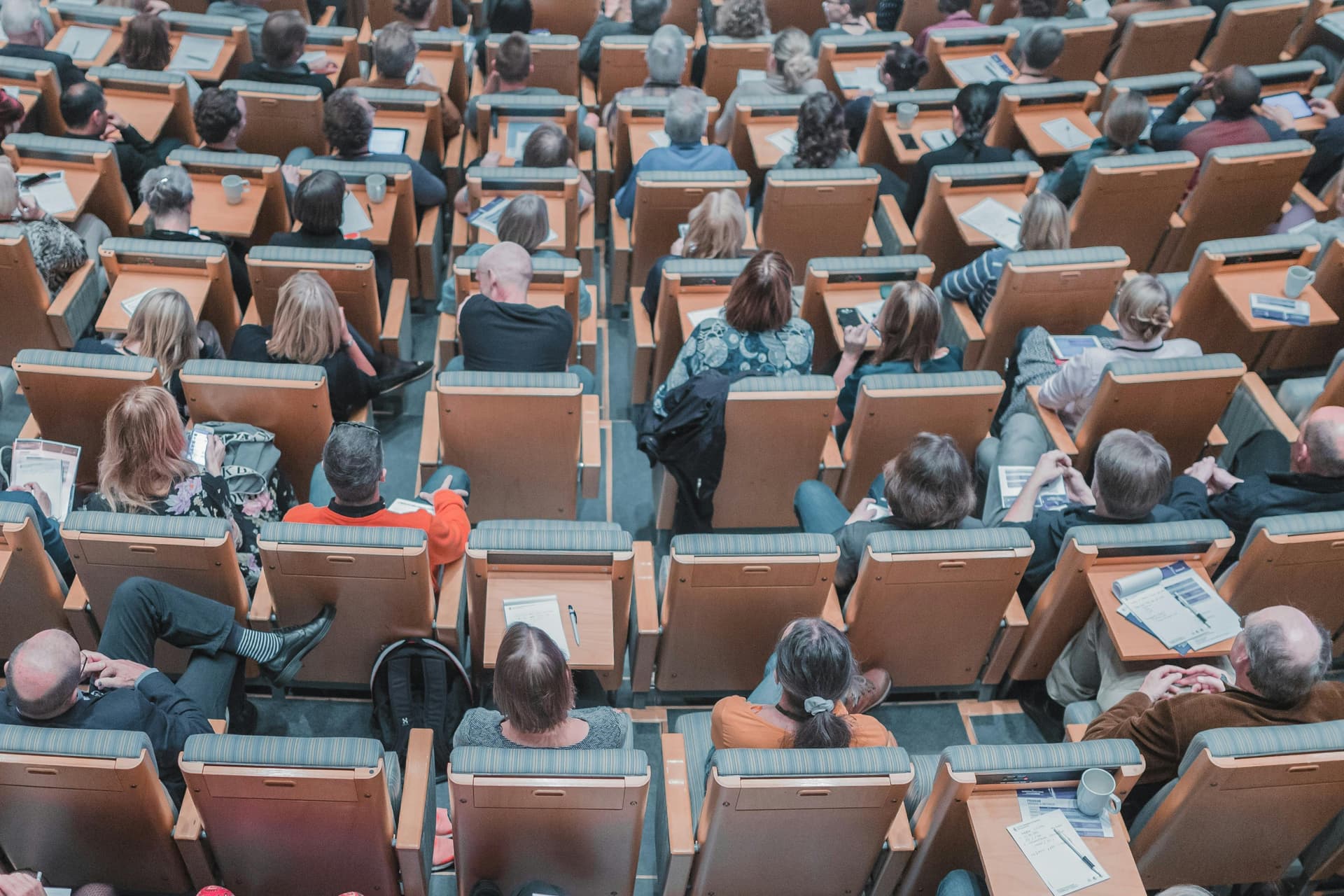 People sitting, reading, and writing in a large auditorium with wooden seats, viewed from above. Some seats have open notepads and cups on their small built-in desks.